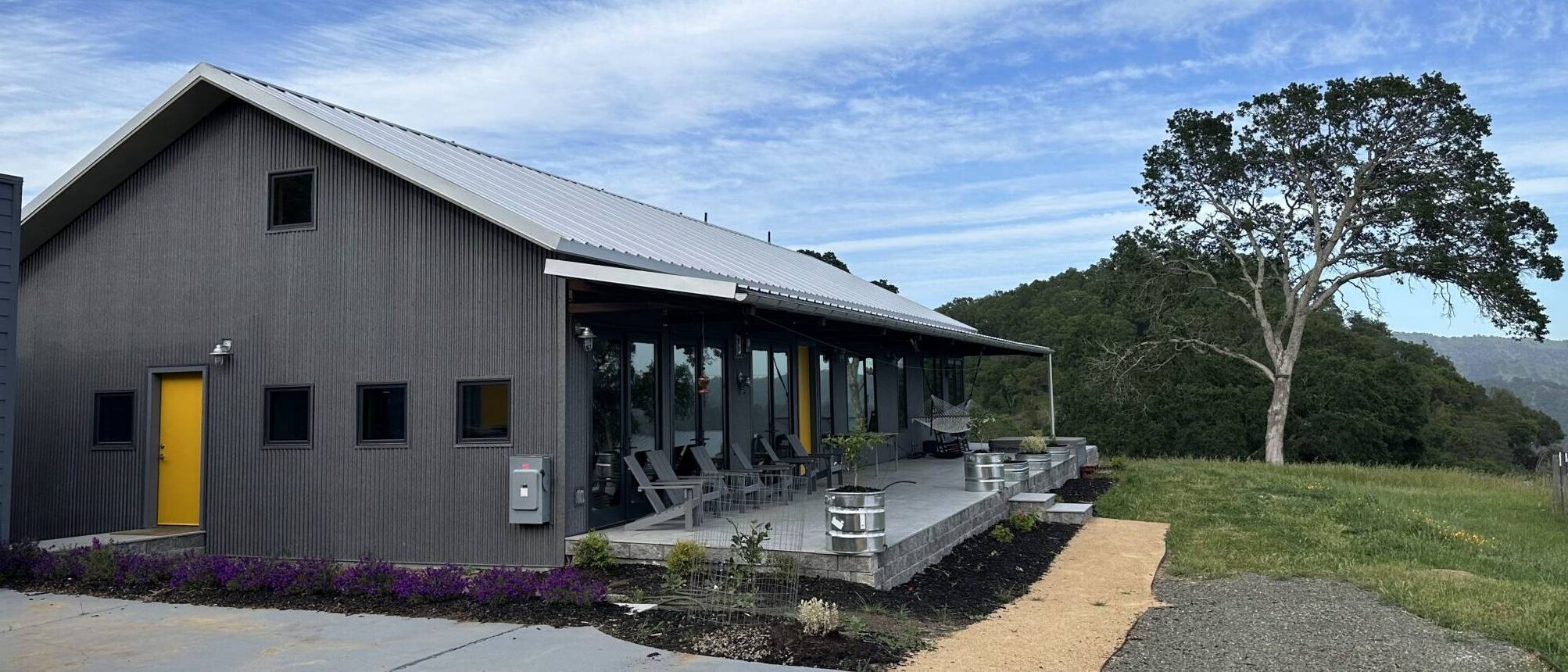 A contemporary building with dark grey corrugated metal siding and a grey metal roof, nestled into a green hillside under a cloudy sky. The building features a bright yellow door on the left and a long row of large windows on the right, which open onto a concrete patio with several lounge chairs and small planters with purple flowers. A large, leafy tree stands on the hillside to the right of the building.