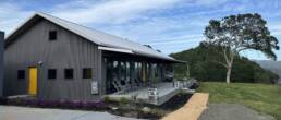 A contemporary building with dark grey corrugated metal siding and a grey metal roof, nestled into a green hillside under a cloudy sky. The building features a bright yellow door on the left and a long row of large windows on the right, which open onto a concrete patio with several lounge chairs and small planters with purple flowers. A large, leafy tree stands on the hillside to the right of the building.