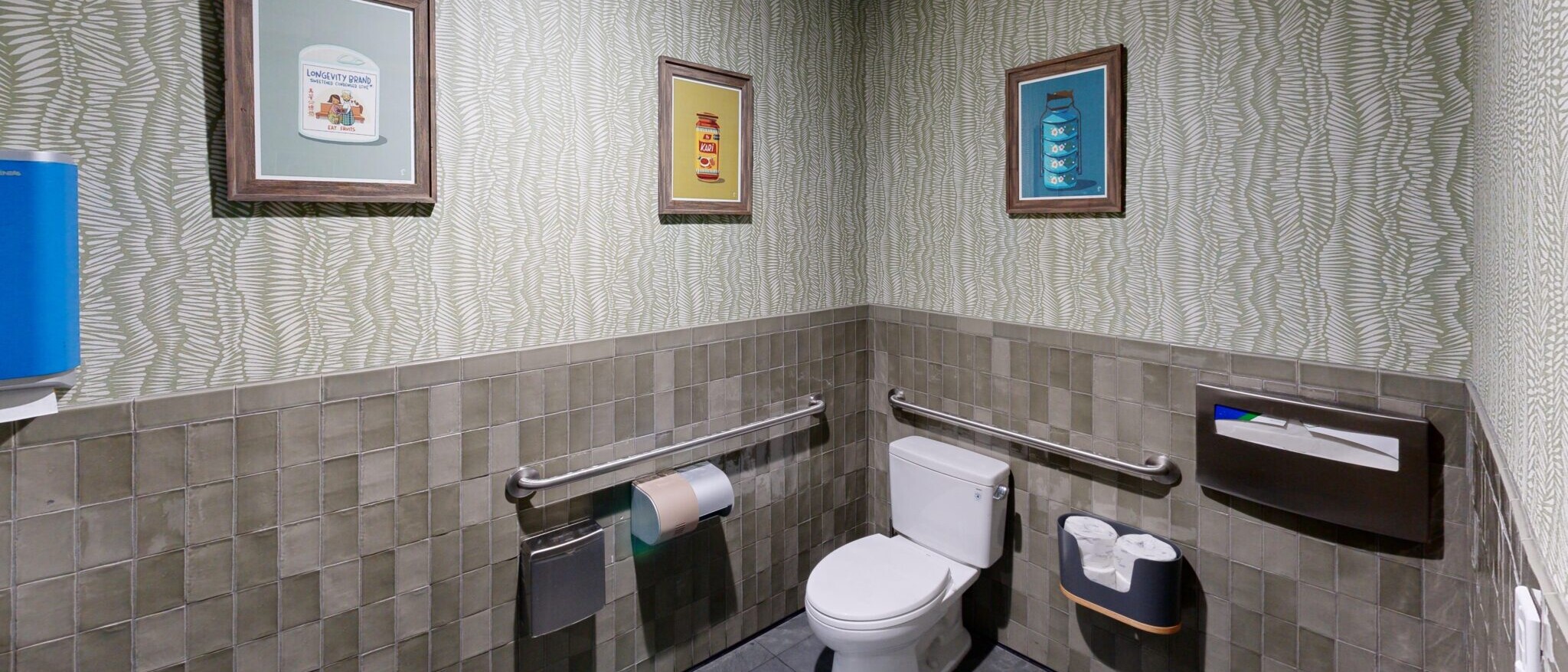 A contemporary public or commercial restroom by Black Creek Builders, featuring distinctive light green textured wallpaper above dark grey subway tiles. The room includes a white toilet with a grab bar on the left, a toilet paper dispenser, and a roll of brown paper towels. Three framed art prints hang on the wall, and a blue dispenser is visible on the left wall. The floor is dark grey tile.