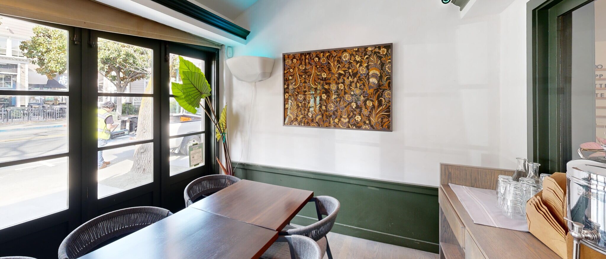 A modern and inviting dining area or cafe space by Black Creek Builders, featuring a long communal wooden table with several grey woven chairs. Large windows with dark frames and light roller shades offer a view to the outside street. The walls are white with a dark green wainscoting, and a textured art piece hangs on the far wall. On the right, a light wood counter serves as a service area with stacked plates and metal containers. Recessed lighting and linear vents are visible in the ceiling.