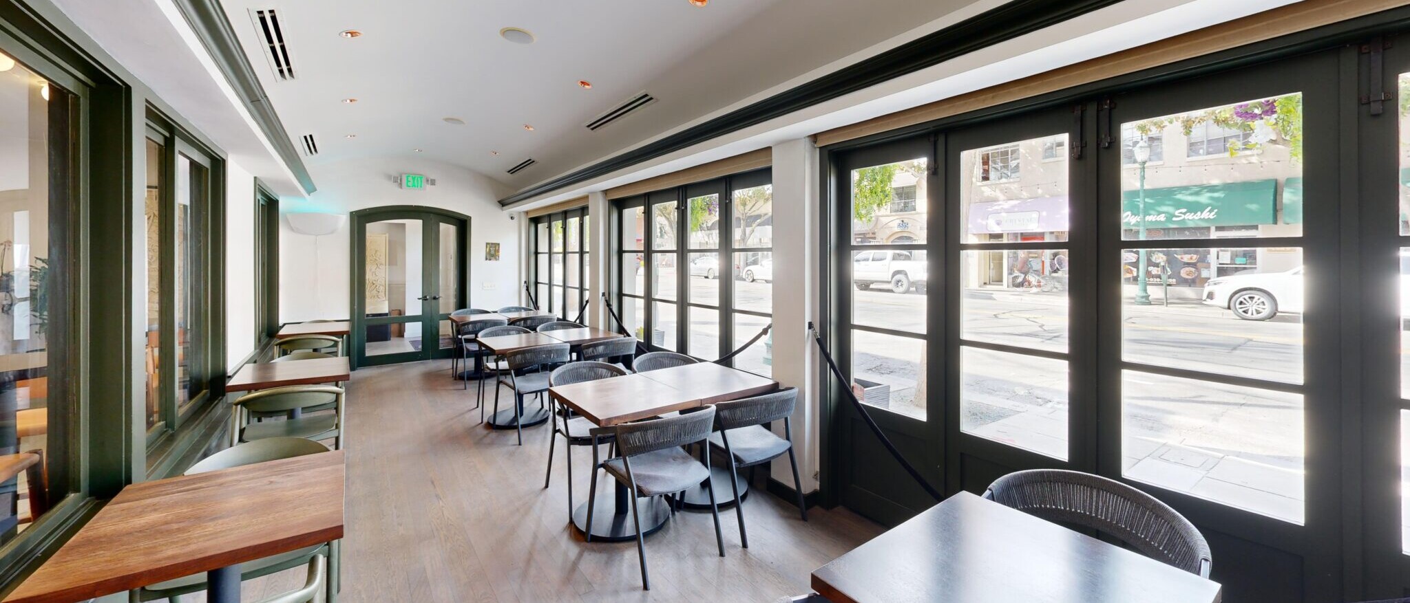 A well-lit restaurant dining area or patio space by Black Creek Builders, featuring numerous dark-framed windows and glass doors that open to a street view. Inside, there are multiple tables with dark wooden tops and light grey chairs, as well as counter-height tables with green chairs along the left wall. The floor is light wood, and the ceiling has recessed lighting and linear vents. A dark green archway is visible at the far end of the room.