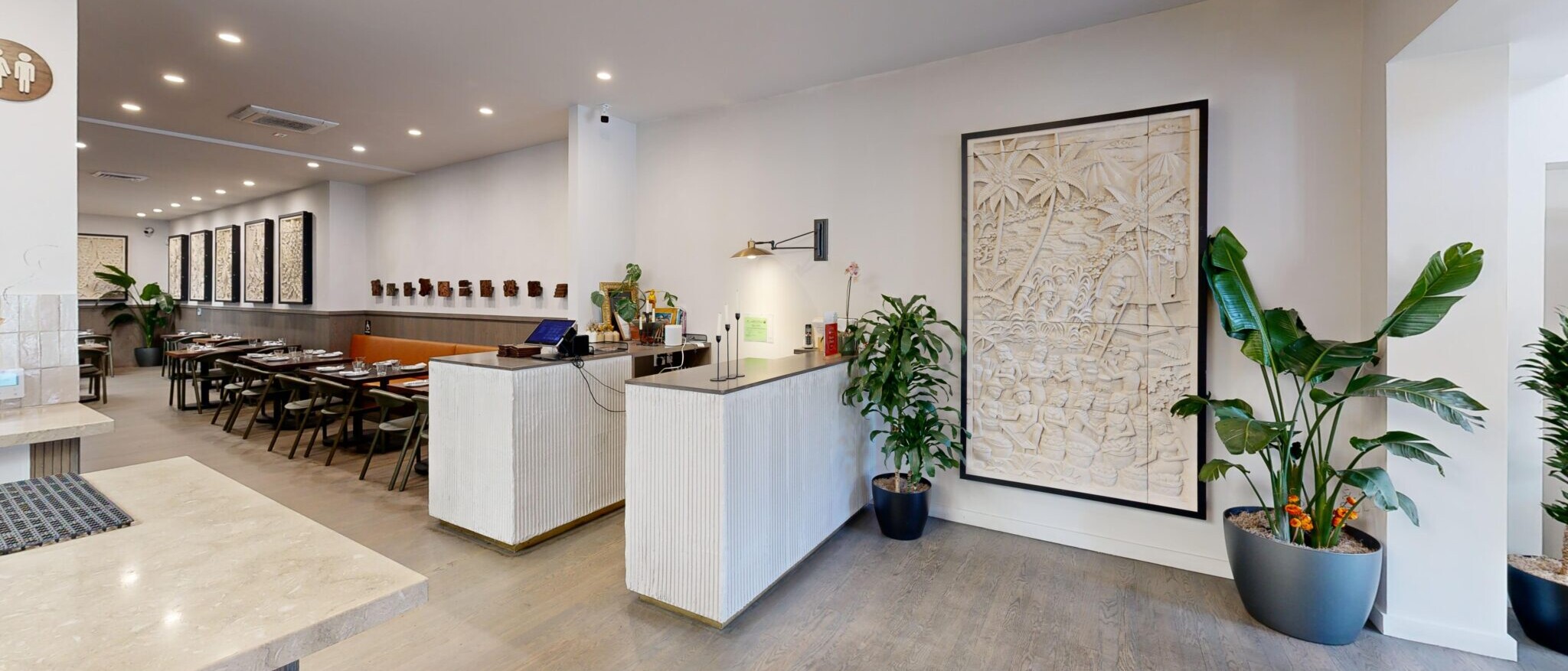 A welcoming and modern restaurant or cafe interior by Black Creek Builders, featuring a spacious layout with a long, fluted white counter serving as a reception or service desk. To the left, a dining area with banquette seating and tables is visible. Large potted green plants, some with orange flowers, are placed throughout the room, including next to a prominent textured wall art piece on the right. The floor is light wood, and the ceiling has ample recessed lighting.