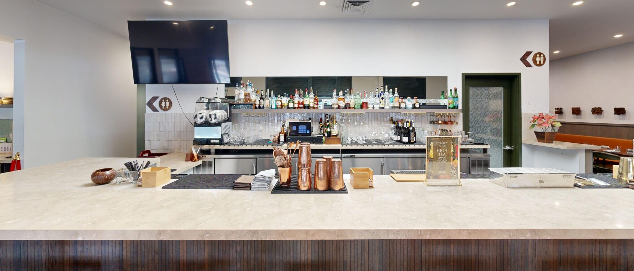 A sophisticated and fully stocked bar area by Black Creek Builders, featuring a long, light-colored marble countertop with brown upholstered barstools in the foreground. Behind the bar, a wide selection of liquor bottles is displayed on shelves with a mirrored backsplash. A large flat-screen TV is mounted on the wall above the bar, and various bar tools and copper mugs are arranged on the counter. Recessed lighting and a large skylight illuminate the space.