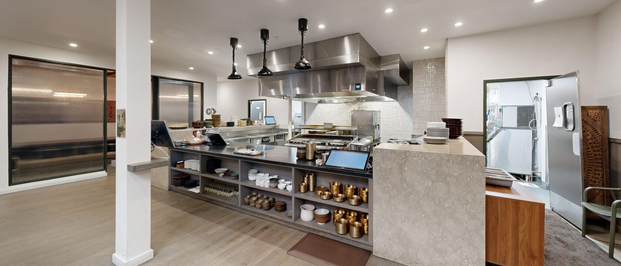 A modern open-concept commercial kitchen by Black Creek Builders, featuring a large stainless steel range hood and professional cooking equipment. In the foreground, a substantial island with open shelving displays various bronze-toned pots and containers, creating an interactive dining experience. To the left, a textured glass partition separates the kitchen from another area. The space is well-lit with recessed lighting and pendant lights over the main cooking area, and has light wood flooring.