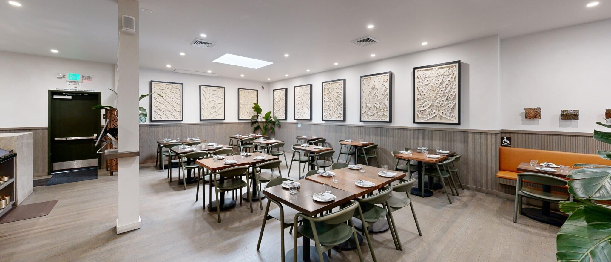 A contemporary and elegantly designed restaurant dining room by Black Creek Builders, featuring numerous wooden tables with dark grey chairs. The walls are adorned with multiple framed abstract art pieces. A light grey wainscoting runs along the bottom half of the walls, complementing the light wood-look flooring. On the right, an orange booth provides additional seating with an accessible table. The ceiling features recessed lighting and a skylight, providing ample illumination.