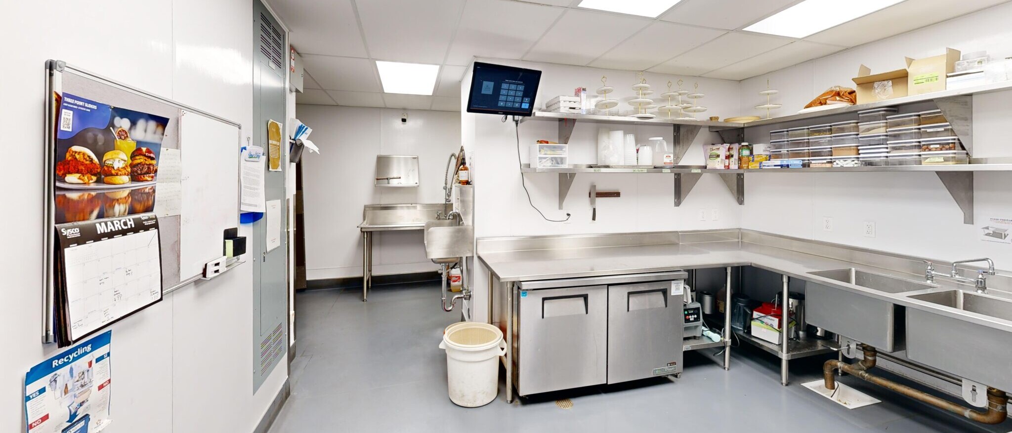 A highly organized and functional catering prep kitchen by Black Creek Builders, featuring white paneled walls and a light grey floor. Stainless steel work tables and counters line the walls, holding stacks of plates, bowls, and other kitchenware. A large stainless steel three-compartment sink is visible on the right. A whiteboard calendar for 