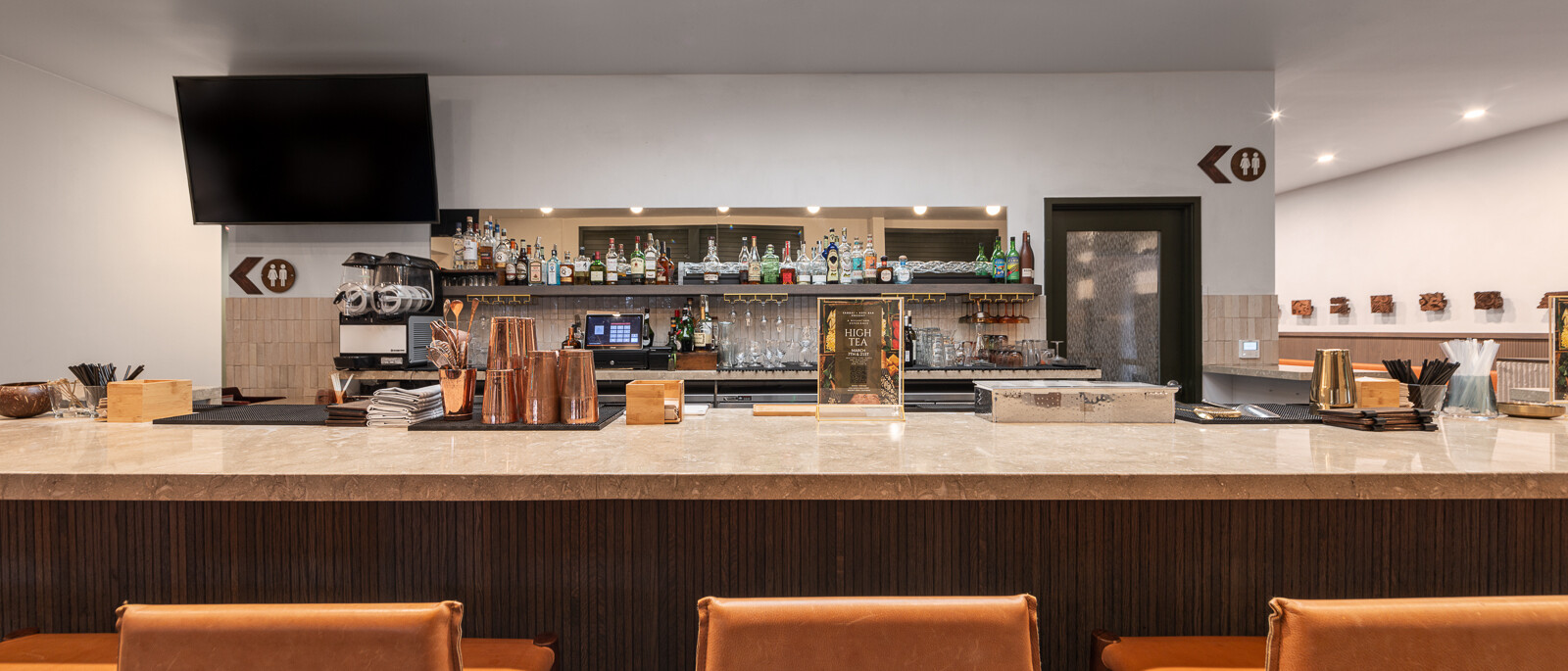 A close-up view of a well-appointed bar area by Black Creek Builders, featuring a light-toned marble countertop and a dark, fluted base. Three light brown upholstered barstools are visible in the foreground. Behind the bar, a variety of liquor bottles and glasses are neatly arranged on shelves against a white wall. A large black flat-screen TV is mounted above the bar on the left, and a large skylight brightens the space from above.