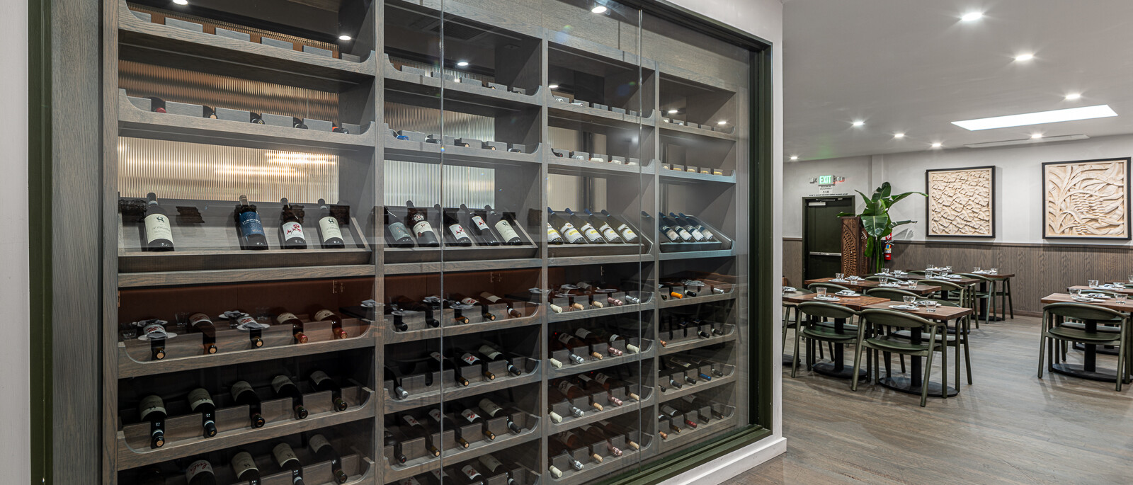 A well-designed, large walk-in wine cellar by Black Creek Builders, enclosed behind a clear glass wall with dark framing. Inside, multiple shelves are neatly stocked with wine bottles, some standing upright and others lying on their sides. The cellar features interior lighting to showcase the collection. To the right, a warm and inviting dining area with wooden tables and chairs is visible, adorned with wall art and illuminated by recessed lighting and a skylight. The flooring is a light wood.