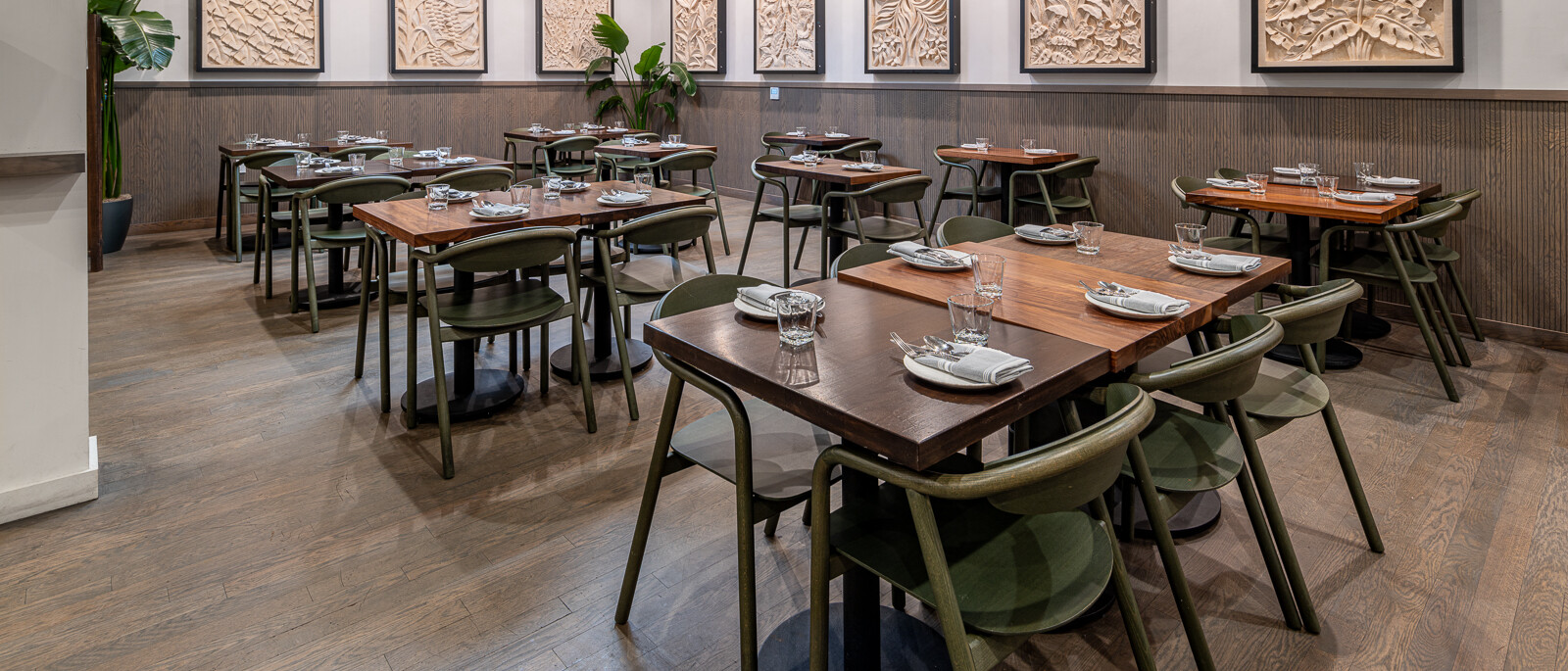 A contemporary and inviting restaurant dining room by Black Creek Builders, featuring numerous wooden tables with dark green chairs. The walls have a light-colored wainscoting and are adorned with multiple framed abstract art pieces in varying sizes. A large skylight and recessed lighting illuminate the space, which has light wood-look flooring. A large potted plant adds greenery to the left side of the room.