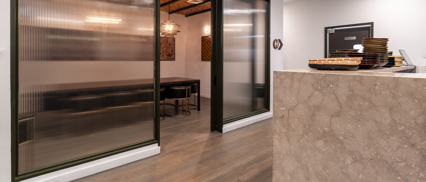 A modern interior space by Black Creek Builders, featuring a large, dark-framed fluted glass sliding door on the left, offering a glimpse into a private dining or meeting room with warm lighting. In the foreground on the right, a light-colored marble reception or service counter is visible, with a display of plates. The room has light wood-look flooring and white walls with recessed lighting.