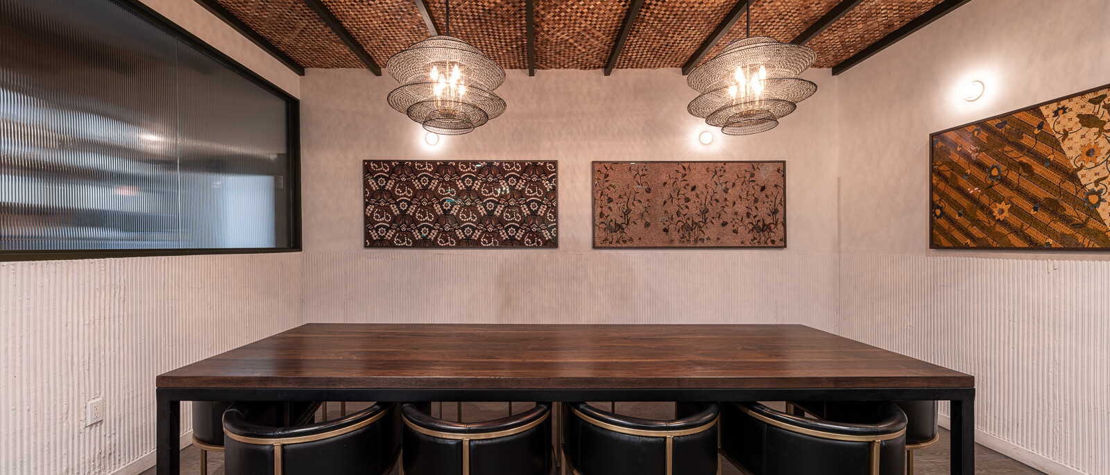 A unique and modern private dining or meeting room by Black Creek Builders, featuring a distinctive woven wood ceiling with exposed beams. Two large, ornate spherical pendant lights hang over a long, dark wooden table with six black chairs featuring gold-toned frames. The walls are light-colored with a textured wainscoting along the bottom, and three framed art pieces hang on the far wall. A window with a horizontal textured glass panel is visible on the left. The flooring is a light wood.