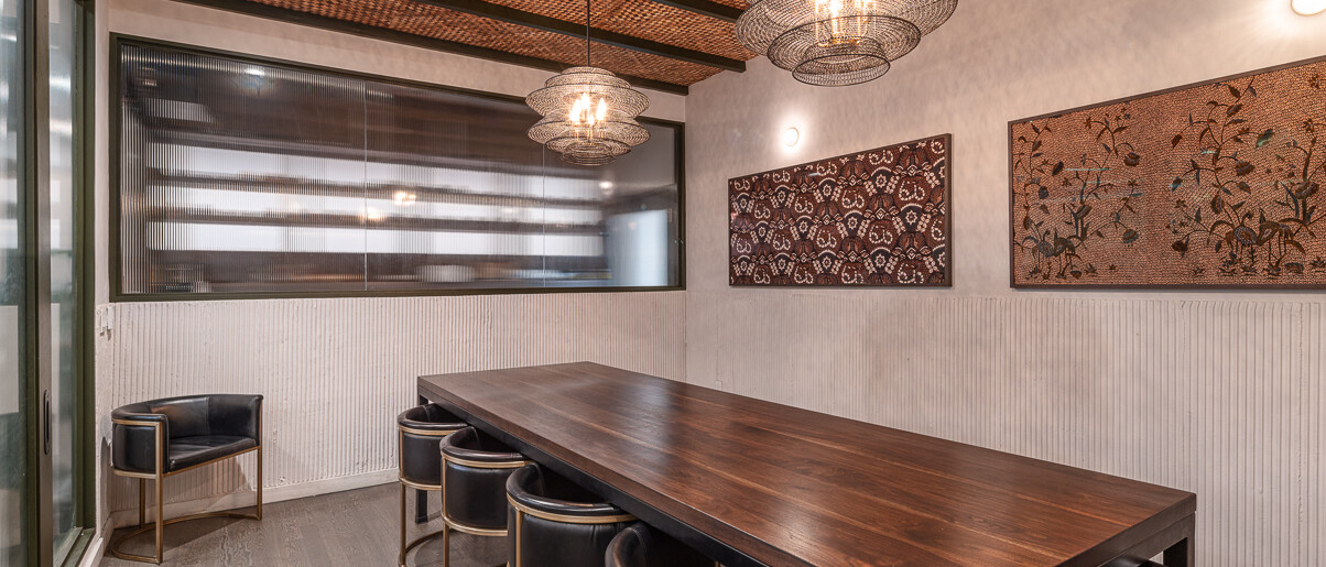 A contemporary private dining or meeting room by Black Creek Builders, featuring a distinctive woven straw ceiling with dark support beams. Two multi-tiered, spherical pendant lights hang over a long, dark wooden table with six black chairs that have elegant gold-toned metal frames. The walls are light-colored with textured wainscoting along the lower half, and two framed abstract art pieces in warm, earthy tones adorn the far wall. A window with horizontal textured glass is visible on the left. The room has light wood-look flooring.