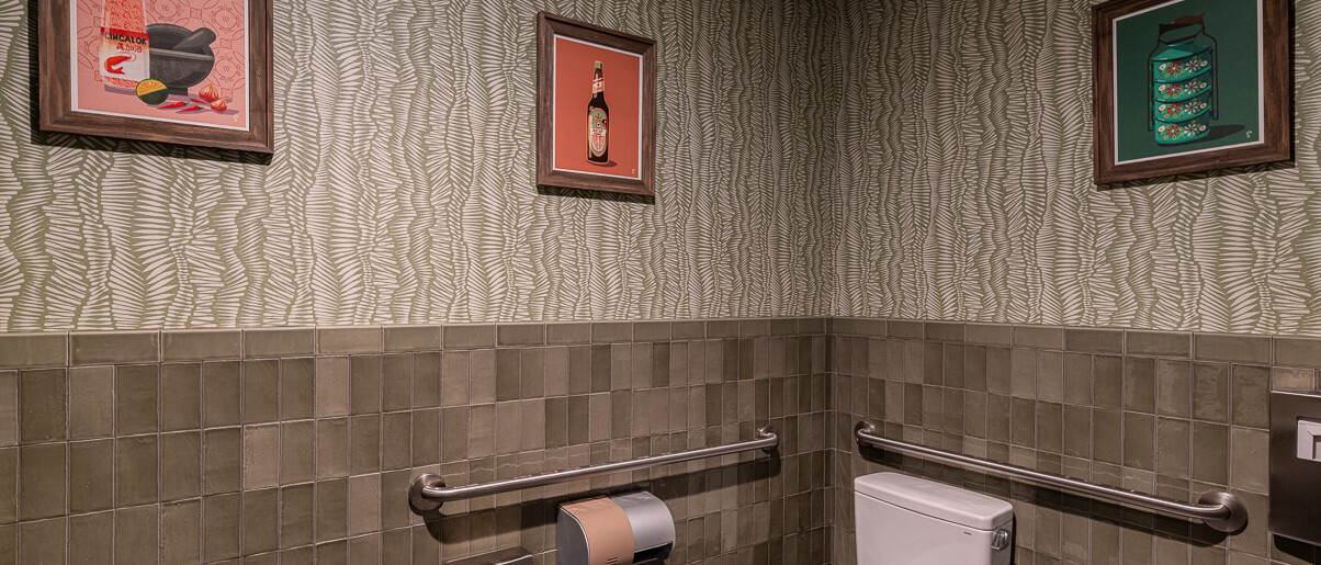 A contemporary and uniquely styled powder room by Black Creek Builders, featuring walls with a wavy light green and white patterned wallpaper above a wainscoting of dark grey rectangular tiles. A white toilet with a grab bar is visible on the right, along with a toilet paper dispenser and a black and orange paper towel dispenser. Three framed art pieces with various graphic designs hang on the walls. The floor is dark grey tile.