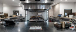 A high-end commercial or luxury residential kitchen by Black Creek Builders, featuring a large central island with a dark countertop and integrated sink in the foreground. Above the island, a massive stainless steel range hood with three black pendant lights is centered over a professional-grade grill or stove. The kitchen has multiple stainless steel work surfaces, stacked plates, and various kitchen tools. White tiled walls are visible in the background, and multiple doorways lead to other areas.
