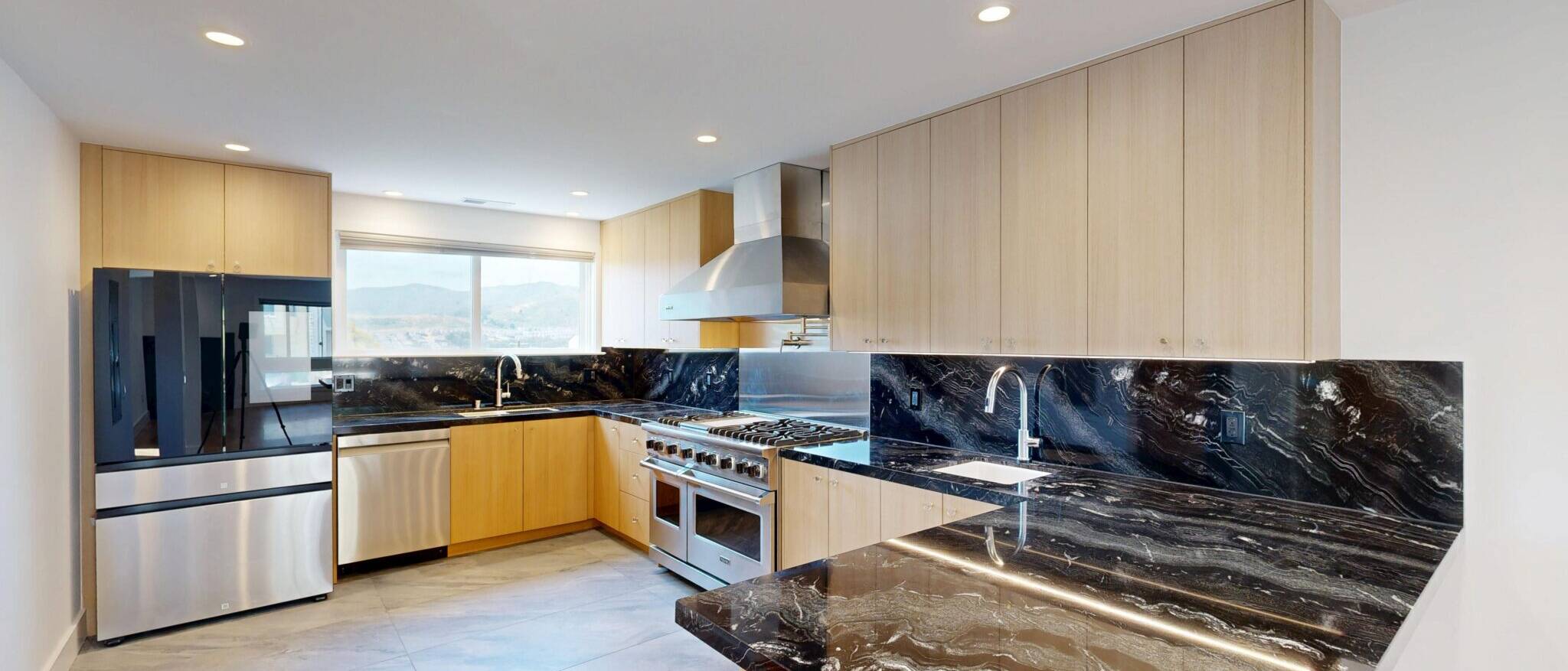 A wide view of a sleek, modern kitchen by Black Creek Builders, featuring black marble countertops with elegant white and gold veining, complementing light wood cabinetry. The kitchen is equipped with stainless steel appliances, including a large refrigerator, dishwasher, professional range, and matching hood. A large window over the sink offers a view of a mountainous landscape. The floor is a light grey tile, and recessed lighting illuminates the space.