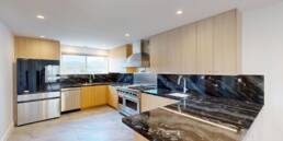 A wide view of a sleek, modern kitchen by Black Creek Builders, featuring black marble countertops with elegant white and gold veining, complementing light wood cabinetry. The kitchen is equipped with stainless steel appliances, including a large refrigerator, dishwasher, professional range, and matching hood. A large window over the sink offers a view of a mountainous landscape. The floor is a light grey tile, and recessed lighting illuminates the space.