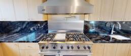 A close-up view of a high-end kitchen by Black Creek Builders, showcasing a professional stainless steel range with a large matching hood. The range is set against a black marble countertop and backsplash with prominent blue veining. Above the range, a wall-mounted pot filler faucet is visible. Light wood cabinetry frames the appliances.