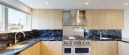 A contemporary kitchen by Black Creek Builders, featuring rich dark blue and black veined marble countertops and backsplash, light natural wood cabinetry, and high-end stainless steel appliances including a professional-grade range and matching hood. A large window above the sink offers a view of a cityscape and hills. Recessed lighting illuminates the space.