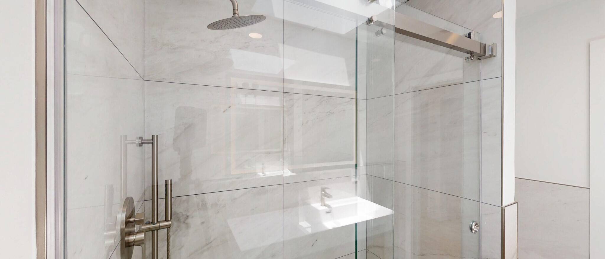 A pristine, modern walk-in shower by Black Creek Builders, featuring large white marble-look tiles on the walls and a clear glass sliding door. The shower is equipped with a brushed nickel rainfall showerhead and a contemporary mixing valve. Reflections on the glass door suggest a bright bathroom environment.