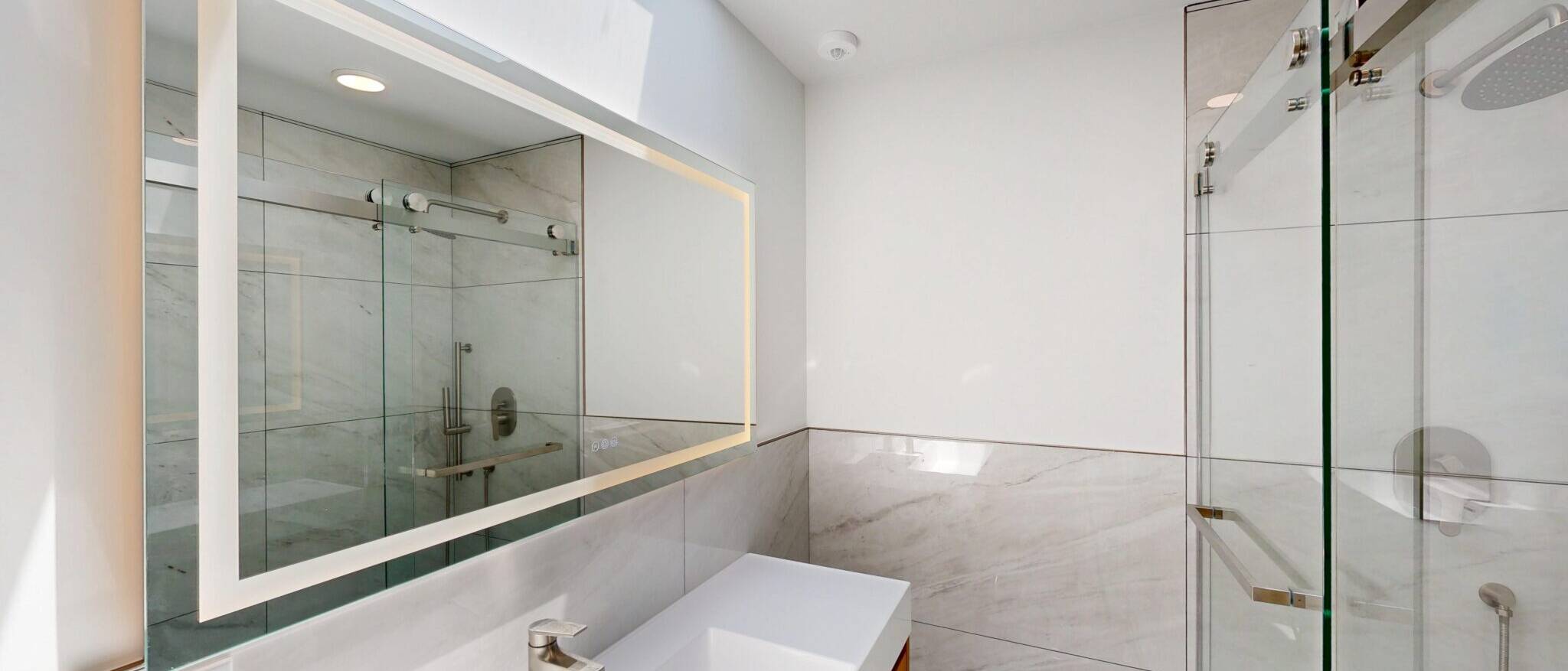 A sleek, modern bathroom by Black Creek Builders, featuring white marble-look tiles on the walls, a large illuminated mirror over a white rectangular sink with a brushed nickel faucet, and a spacious glass-enclosed shower on the right. The room is brightly lit with recessed lighting and natural light from above.
