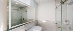 A sleek, modern bathroom by Black Creek Builders, featuring white marble-look tiles on the walls, a large illuminated mirror over a white rectangular sink with a brushed nickel faucet, and a spacious glass-enclosed shower on the right. The room is brightly lit with recessed lighting and natural light from above.