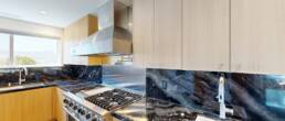 A luxurious modern kitchen by Black Creek Builders, featuring striking black and blue veined marble countertops and backsplash, light wood cabinetry, and stainless steel appliances including a large range hood and a professional-grade stove. A window offers natural light and a view in the background.