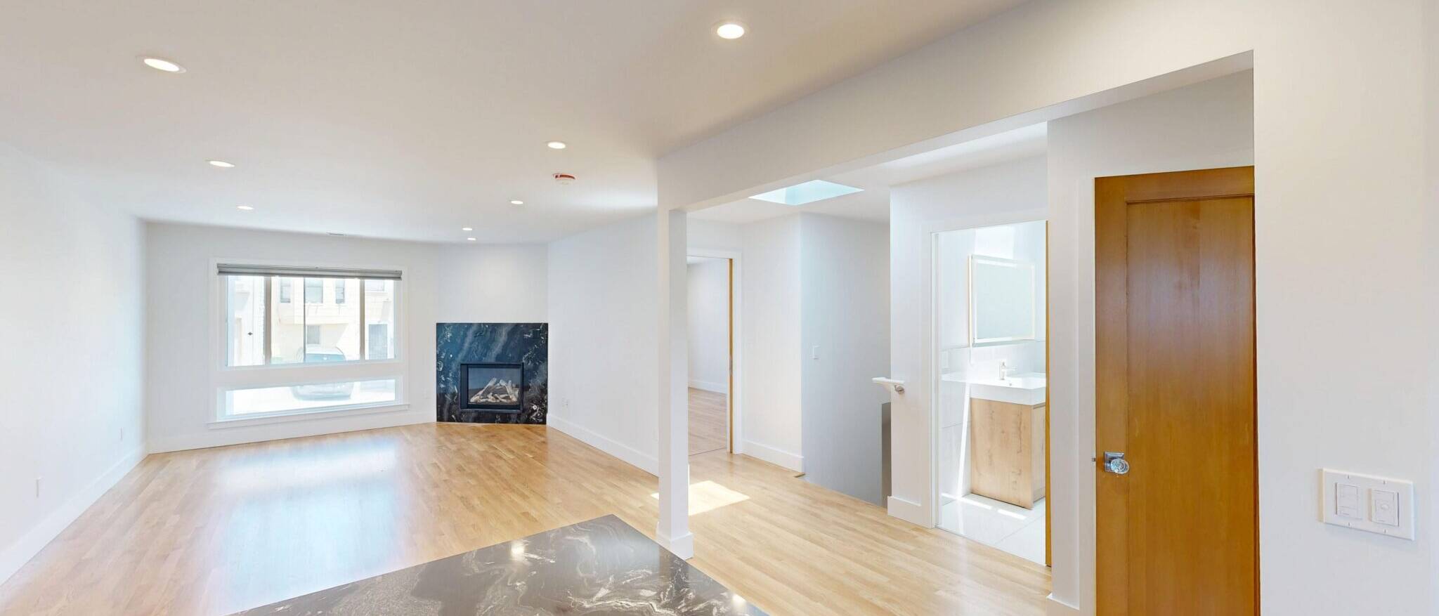A modern, open-concept living space by Black Creek Builders, featuring light hardwood floors and white walls. In the foreground, a dark marble-patterned countertop is visible. The living area includes a black marble fireplace and a large window. Beyond, a wooden door leads to another room, and a bathroom vanity can be seen in the distance.